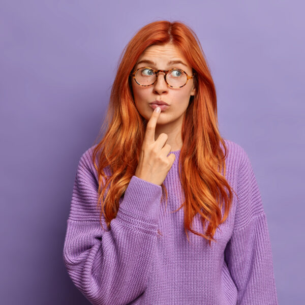 Portrairt of thoughtful ginger woman looks pensively aside, keeps index finger on lips and wears warm sweater. Beautiful blue eyed lady with red hair poses over purple background, ponders on something